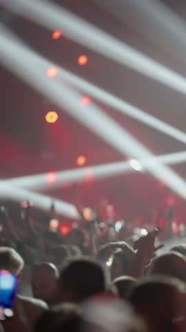 Slow-motion video from a crowd at a live music concert. People raise their hands and film with phones as dynamic stage lights flash. Energetic, immersive atmosphere. Vertical format.