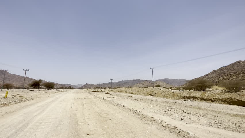 A wide-angle view of a dirt road winding through a vast, arid mountain landscape featuring utility lines and sparse desert vegetation under a clear sky.