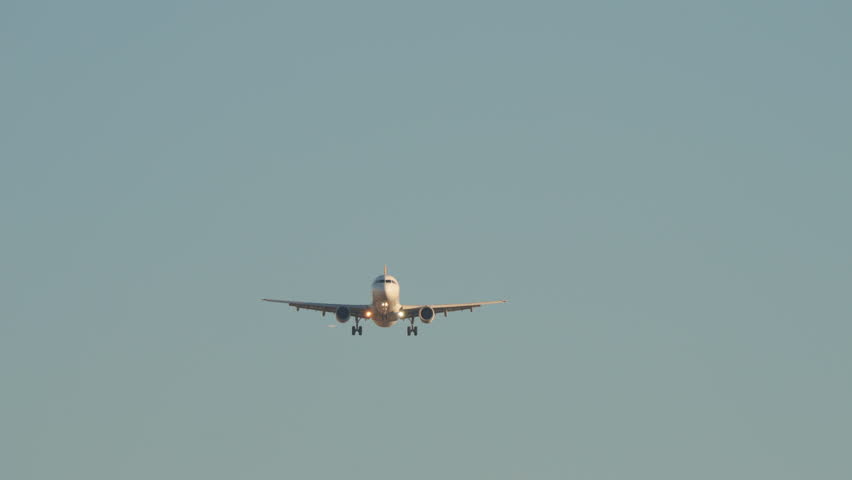 Video footage shows a commercial airplane on final approach. The jetliner flies directly towards the camera and then passes low overhead, showing its undercarriage and landing gear. Clear blue sky.