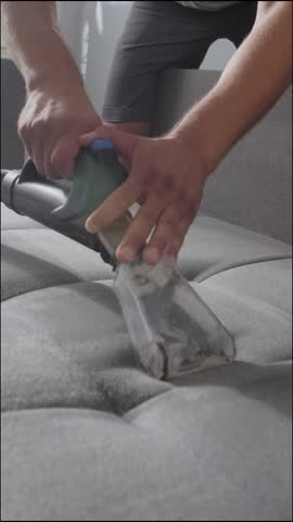 Close-up of a worker cleaning a dirty grey sofa with a professional wet vacuum cleaner