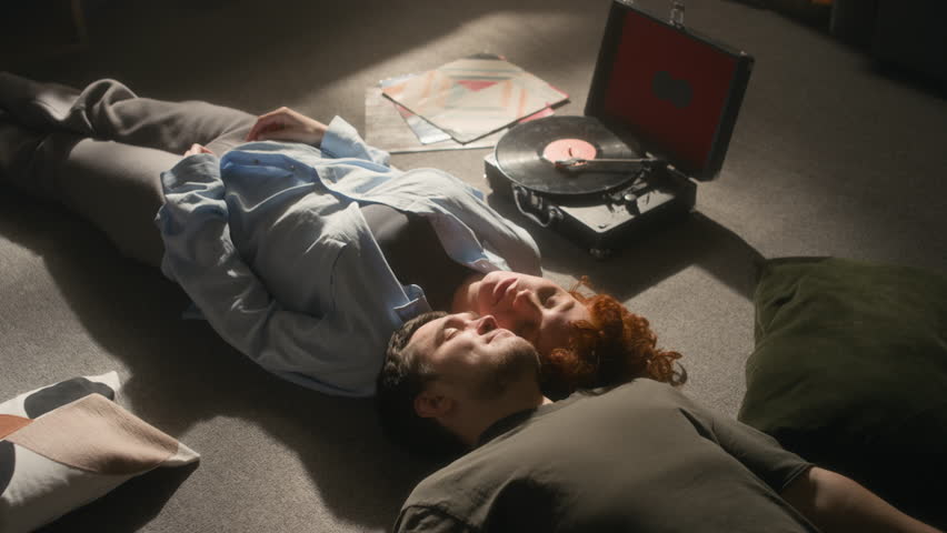 Cinematic high angle shot of happy young couple lying on floor with eyes closed and listening to music with retro record player in serene sunlight, copy space