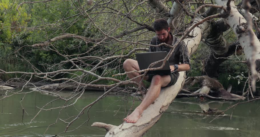 Man using laptop outdoors doing remote work replying emails in nature, sitting on a fallen tree in a wild river