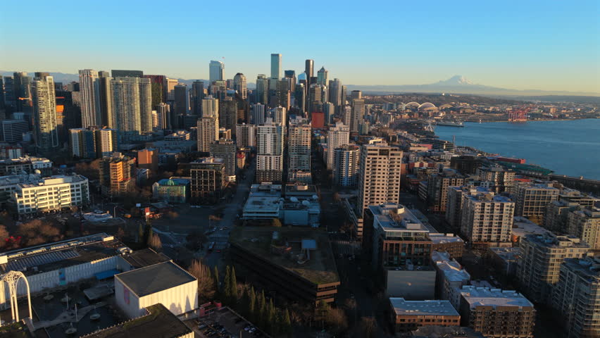 Aerial cityscape of Seattle Washington downtown with modern skyscrapers, residential towers and Puget Sound coastline during golden hour, showing urban density and calm evening light.