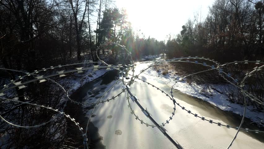 Barbed Wire Fence Over Frozen River In Winter Forest, Sharp Razor Wire Security Barrier. Metal Obstacle In Restricted Area. protective Military Fencing. Border Control And Safety Perimeter Steel Coils