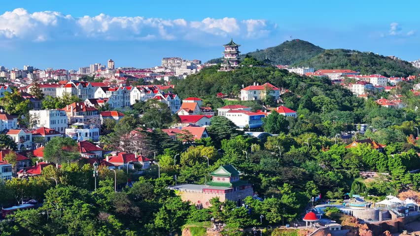 Aerial view of a vibrant hillside city with colorful red-roofed buildings, lush green vegetation, and a traditional pagoda tower overlooking the urban landscape under clear blue sky.