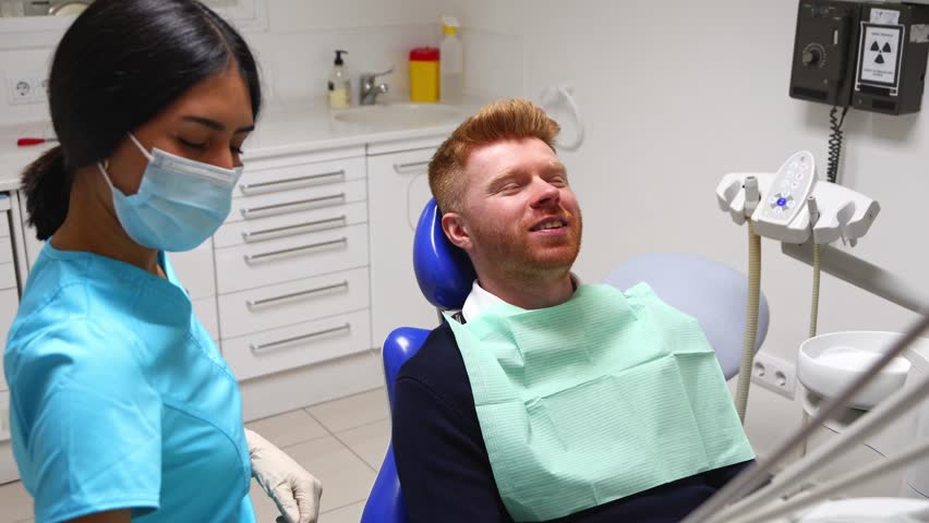 Young man with red hair sitting in a dental chair during a routine checkup. Female dentist in a mask and gloves using tools for oral examination