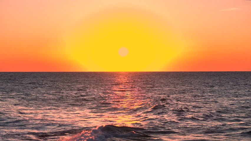 AERIAL, LENS FLARE: Golden setting sun is low over the Mediterranean horizon as small waves roll against a rugged rocky shoreline, catching warm reflections on sea surface. Peaceful scenery at Sicily.