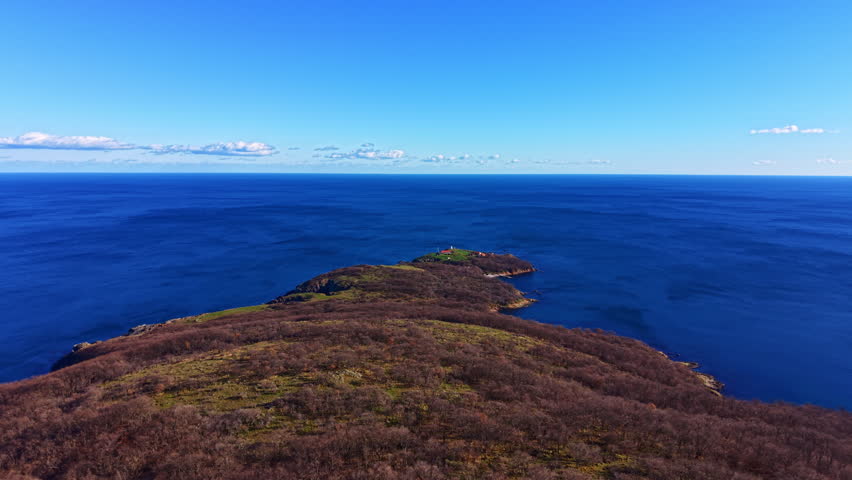 A high view captures a green island with low vegetation surrounded by deep blue water. The sky is clear with a few clouds visible. This scene is located near the coast.