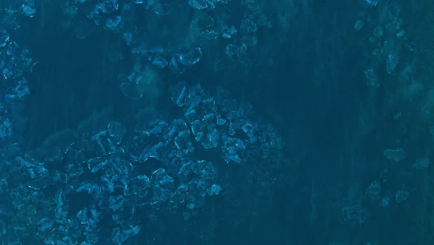 Drone top-down view of water surface during extreme winter cold. Subzero temperatures create floating ice formations. Wind moves small ice fragments across surface.