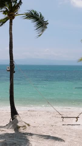 A swing hanging between two palm trees on a white sand beach with turquoise water and clear blue sky. Haad Yuan Beach. Ko Pha Ngan, Thailand.