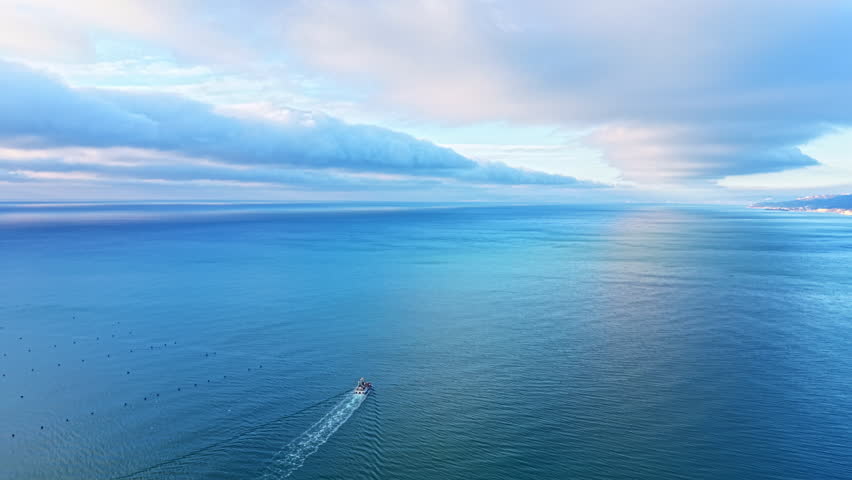 A small boat moves across the vast ocean water during early morning hours. The sky is filled with clouds reflecting light over the blue water. The scenery is peaceful and natural.