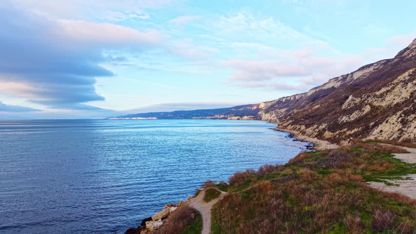 Waves crash against rocks while hills rise above the coastline. The sky has clouds scattered among blue.