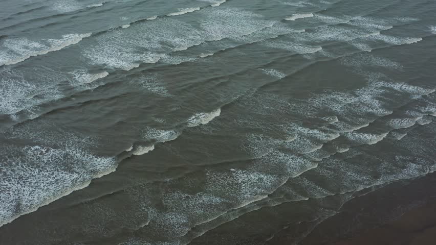 Drone glides over textured grey waves surging toward shore at Talsari Beach under cloudy skies highlighting foam patterns and sea motion.