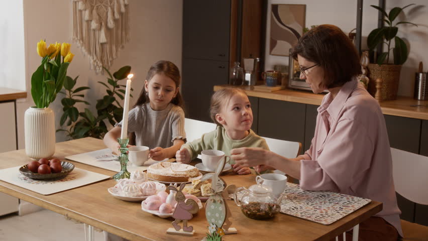 Medium shot of adorable Caucasian little girls celebrating Easter with their loving grandmother, sitting at festive table in cozy modern kitchen and picking dyed eggs