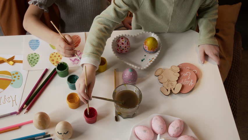 High angle cropped shot of hands of unrecognizable little girls dipping brushes in gouache paint and drawing pattern on dyed eggs while preparing Easter decorations at table