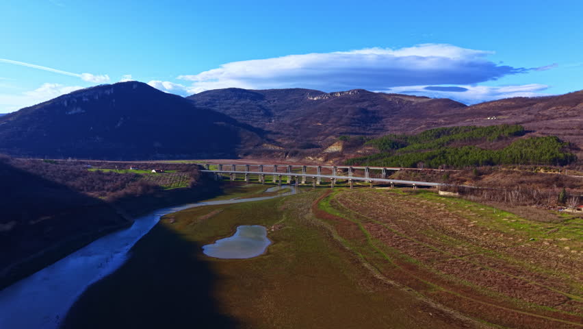 A river runs alongside a bridge that crosses it. Hills rise in the background under a clear blue sky. The landscape shows dry areas and some green patches.