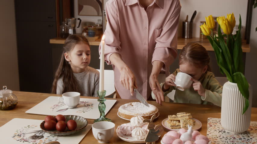 Midsection shot of unrecognizable adult woman cutting cake while celebrating Easter with little girls gathered around festive table decorated with candle, bunny rabbits and dyed eggs