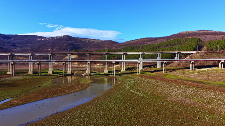 A long bridge stands over a riverbed with low water levels. The ground shows cracked earth and sparse vegetation. Trees and hills provide a backdrop on a bright sunny day.