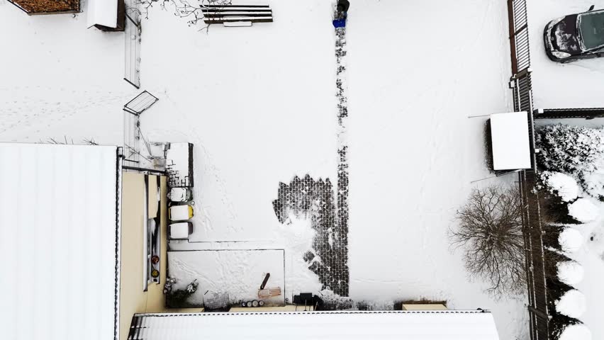 Overhead aerial view of a man clearing snow inside an enclosed urban courtyard, exposing a narrow brick path amid surrounding buildings, trees, and winter-covered surfaces.