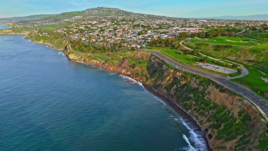 High aerial flyover of stunning San Pedro, California coast at golden hour with beautiful homes above the rugged cliffs, and waves crashing along the shore