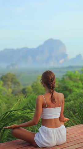 Morning yoga meditation on wooden terrace as woman stretches in lotus pose, overlooking lush jungle and misty mountains in Krabi, Thailand. Ideal for wellness, travel, mindfulness and spa ads. 