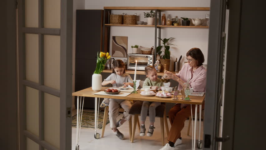 Full shot of caring Caucasian grandmother pouring tea for her adorable little granddaughters gathered around festive table and celebrating Easter together