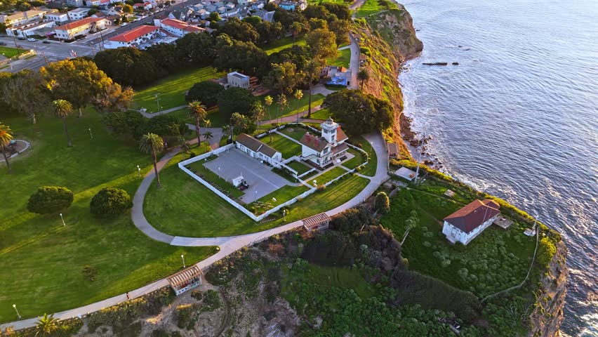 Aerial crane shot of Point Fermin Lighthouse in Los Angeles, California at sunrise with colorful sky, rugged cliffs, and waves crashing along the coast