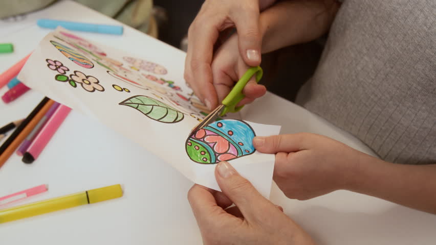 Cropped slowmo shot of hands of unrecognizable adult helping little child to cut out colored Easter egg drawing, engaged in creative activity together