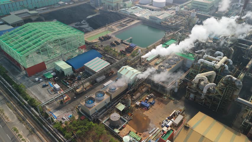 An expansive aerial shot captures the bustling activity of a modern industrial complex, likely a power plant, with numerous buildings, intricate networks of pipes, and prominent cooling towers emitting plumes of steam into the daytime sky. Large piles of coal are visible alongside various water treatment ponds with greenish water, indicating heavy industrial processes. The scene showcases a significant infrastructure dedicated to energy production and material processing, set against a backdrop of distant city buildings and surrounding natural elements. Concept of heavy industry and environmental impact.