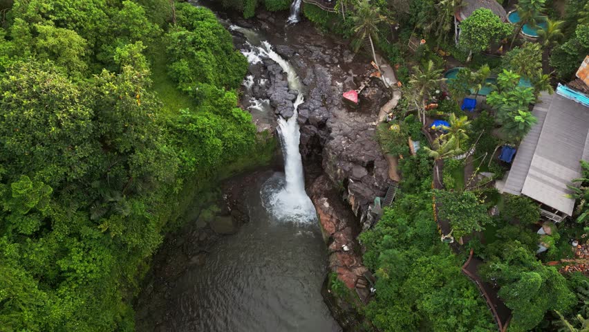 Drone footage revealing a Bali resort with pools located next to a jungle waterfall and river system framed by dense tropical rainforest vegetation, rocky ground and natural forest landscape.