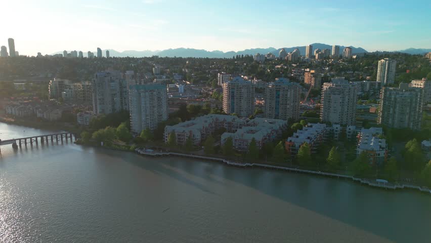 Golden hour drone view of New Westminster showing residential high-rise buildings along the Fraser River with distant mountains in British Columbia, Canada.
