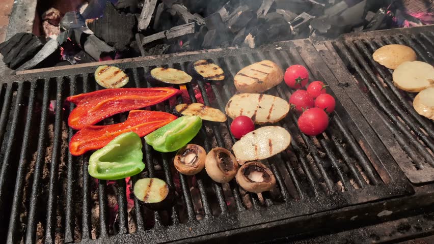 Assortment of delicious fresh vegetables grilling on a hot barbecue over flaming charcoal embers, including red peppers, green peppers, mushrooms, potatoes, zucchini and cherry tomatoes