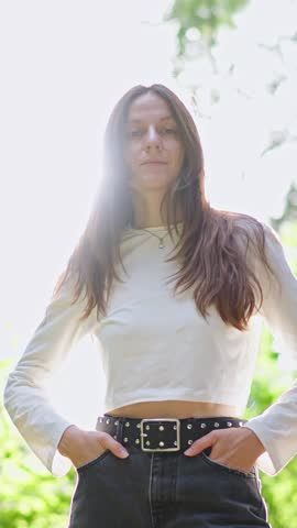 VERTICAL VIDEO: Beautiful young woman smiles calmly and confidently at the camera, posing stylishly with hands in pockets in a sunlit forest.