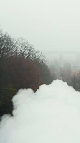 A vintage steam train, emitting thick white smoke, travels along a foggy, rural railway. The train