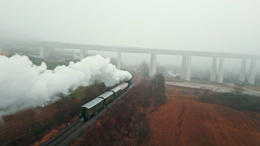 A steam train emits thick white smoke as it travels along a railway track, set against a foggy backdrop with a large bridge overhead. The scene captures a nostalgic, retro style.