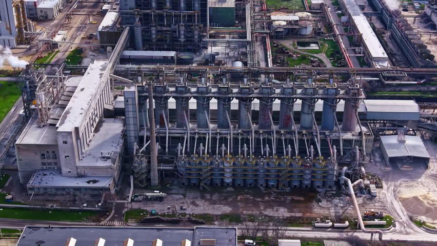 Large industrial site shows multiple silos and machinery working in the background. Nearby buildings and structures are visible. Sky is clear, and daytime setting is noted.