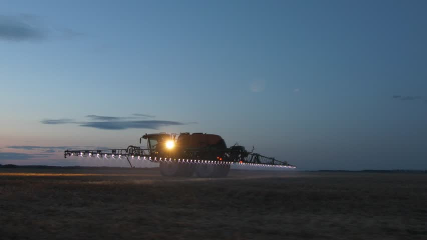 A self-propelled sprayer moves across the field at dusk, illuminating the working area with bright headlights and boom lights. Night spraying allows agricultural operations to be carried out more efficiently by reducing evaporation and improving application accuracy. A calm atmosphere of modern technology and precision farming.