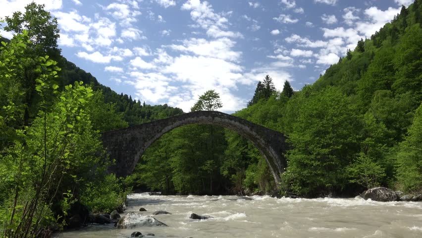 Time lapse of old historical stone arch bridge over rushing river in dense mixed forest of Firtina Valley. Lush Black Sea nature surrounds the ancient bridge in Rize Camlihemsin Turkey.