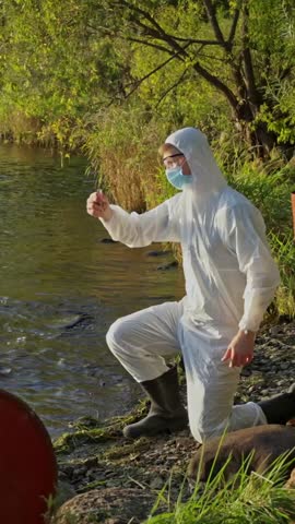 Scientist in hazmat suit looking at the water sample from the river in the test tube, vertical shot