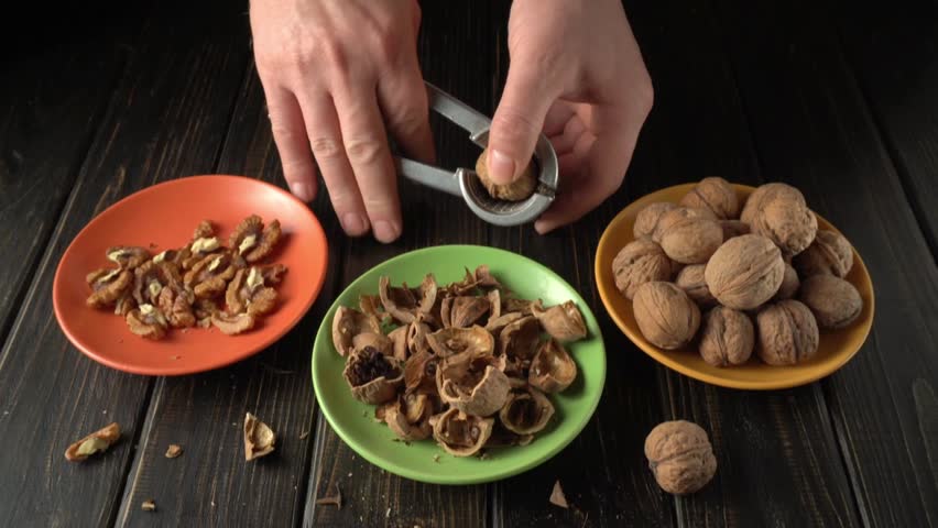 Hands using a nutcracker to crack open walnuts, revealing the nut inside, with empty walnut shells and shelled nuts arranged on colorful plates in a well-lit kitchen setting