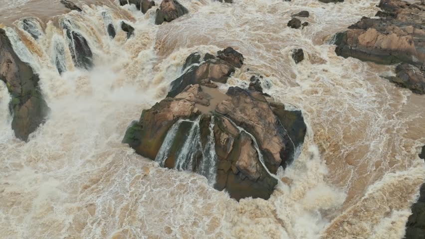 Close aerial view of muddy water plunging over dark rocks forming multiple small cascades at Khone Phapheng Waterfall.