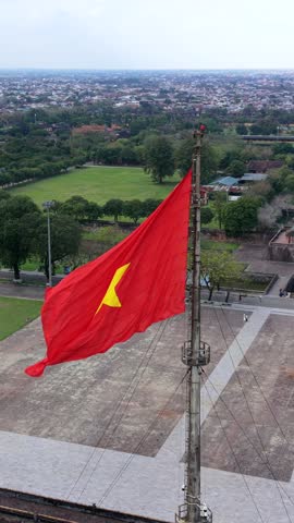 Aerial view of the Vietnamese flag flying above the Imperial City of Hue, Vietnam, with historic palace architecture, city walls, and green gardens visible in the background.