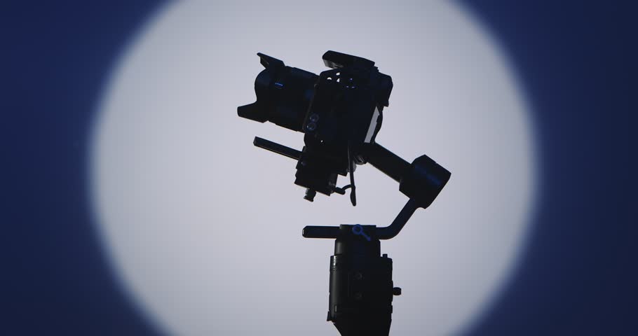 The operator stabilizes the camera mounted on a motorized stabilizer with a suspension, filmed against a bright round light on a dark blue background, symbolizing film production.
