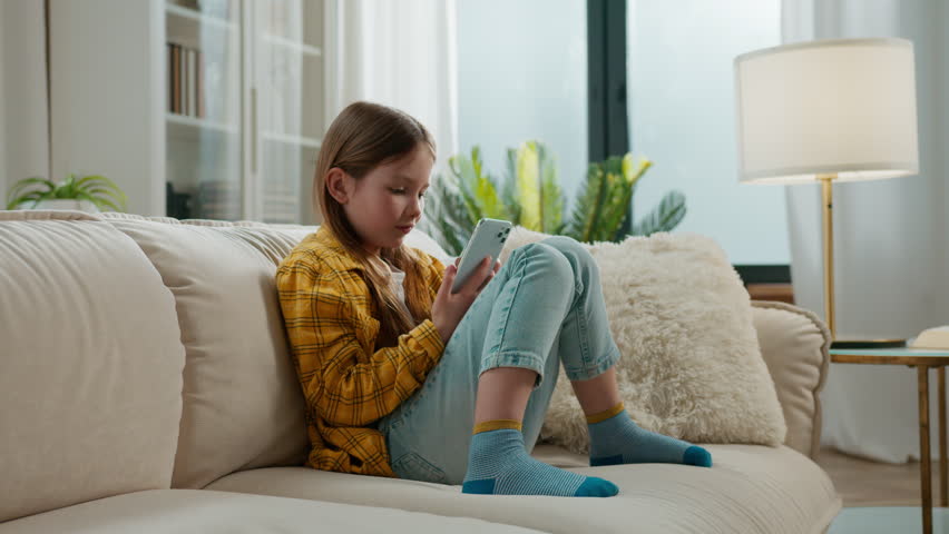 Gadget addict Caucasian little child girl using mobile phone at home kid happy smiling scrolling smartphone technology relaxing on sofa daughter watching funny video in social media smile in apartment