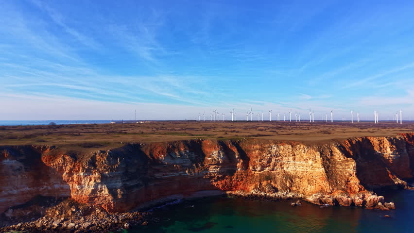 Cliffs rise above the ocean with clear blue water below. Wind turbines line the horizon, standing tall against the blue sky during daytime near the coast.