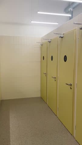 public restroom with yellow stalls and tiled walls, row of closed cubicles with circular windows, metal handles and locks, bright fluorescent lighting, clean Video taken on mobile device.