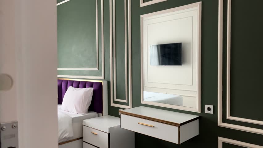 Stylish modern bedroom interior featuring a white bed, purple upholstered headboard, classic green wall panels, minimalist furniture, and a wall-mounted TV with soft natural light.