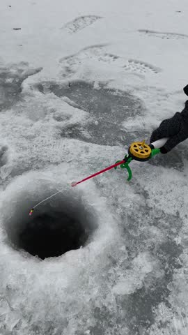 A close-up shot captures the moment of ice fishing on a frozen lake. Snow blankets the icy surface, with a clear hole for fishing. A gloved hand holds an ice fishing tool, featuring a red line and vib