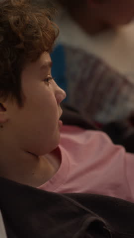 Vertical shot of relaxed preteen boy holding joystick while playing co-op videogame with best friend while hanging out together at home