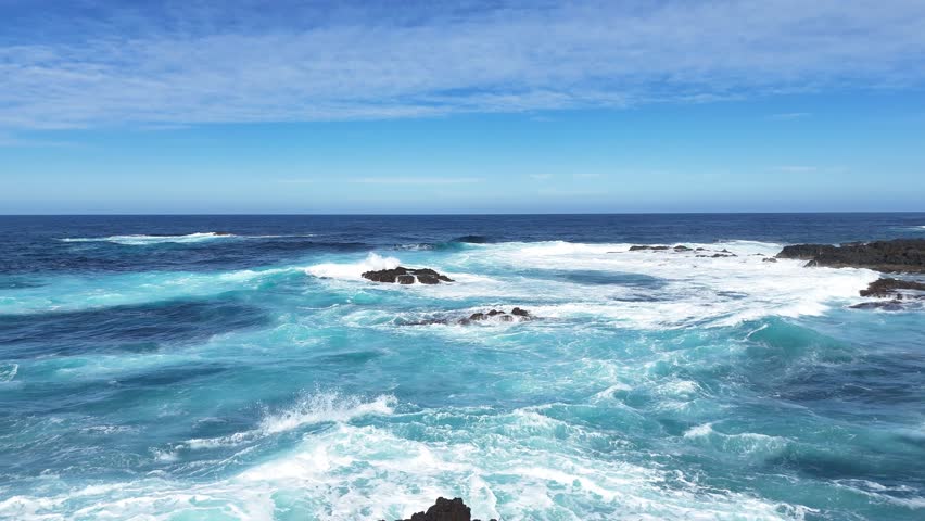 Flying over ocean waves crashing over rocky coastline. Dynamic movement of water and foam against the rugged stone surface under clear blue sky. Aerial view of bright colorful nature. Scenic turquoise